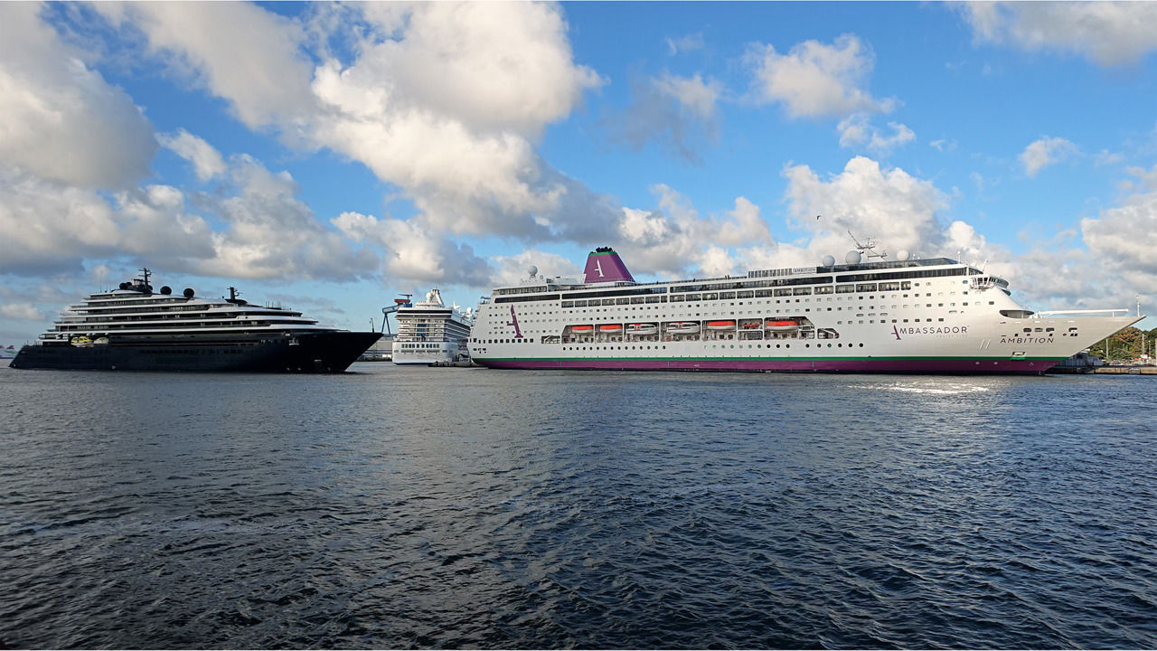 Drei Kreuzfahrtschiffe im Warnemünder Kreuzfahrthafen: links die dunkel gerumpfte Evrima, dahinter die weiße Marina, rechts die Ambition der Ambassador Cruise Line mit magentafarbenem Schornstein und grünem Wasserpass.