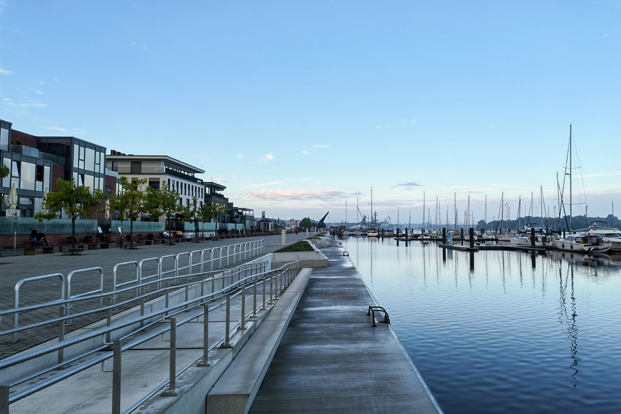 Leere, regennasse Promenade aus Beton direkt am Wasser, links davon Stufen mit Edelstahlgeländern und darüber der gepflasterte Platz vor einer Reihe von Restaurants und Gebäuden mit großen Glasfassaden. Rechts liegen Segelboote an den Stegen der Marina, ihre Masten spiegeln sich im ruhigen Wasser. In der Bildmitte ist in der Ferne der historische Holztretkran zu sehen, darüber ein hellblauer Morgenhimmel.