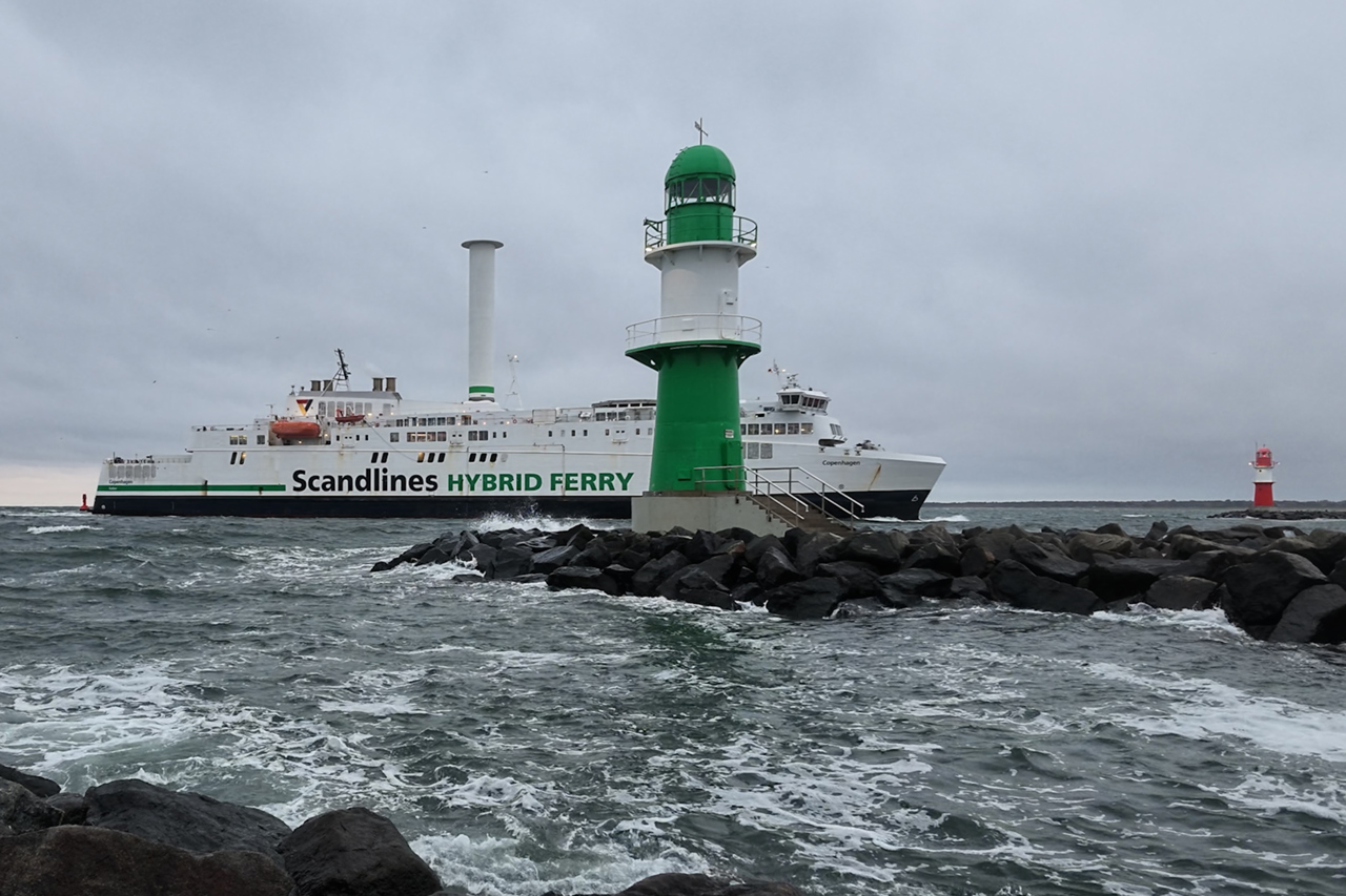 Die Scandlines-Hybridfähre „Copenhagen“ mit Rotorsegel läuft bei aufgewühlter See am grünen Molenfeuer der Westmole vorbei, rechts im Hintergrund das rote Molenfeuer der Ostmole.