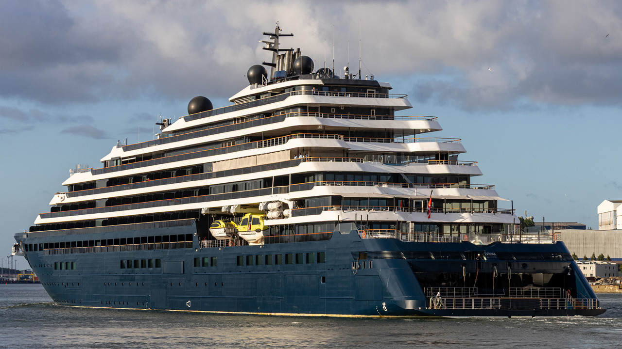 Heckansicht der Evrima im Morgenlicht mit der offenen Marina-Plattform am Heck, den terrassenförmig gestaffelten Balkondecks und den gelben Rettungsbooten.
