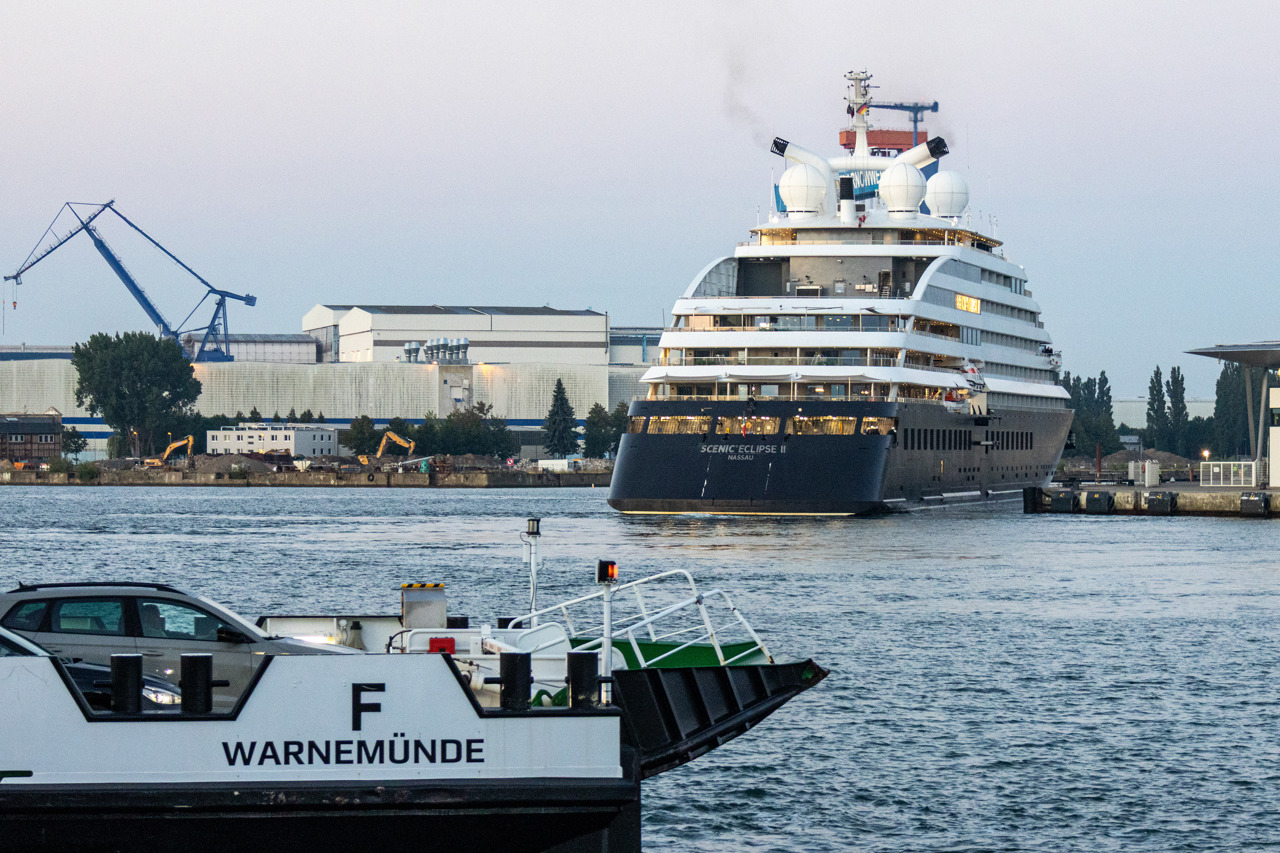 Das dunkelblau-weiße Luxus-Kreuzfahrtschiff „Scenic Eclipse II" mit Heimathafen Nassau bei der Einfahrt in den Warnemünder Hafen in der Morgendämmerung, im Vordergrund der Bug der Fähre „Breitling" mit der Aufschrift „F Warnemünde", im Hintergrund ein blauer Kranausleger und Hallen der Werft, rechts das WCC8