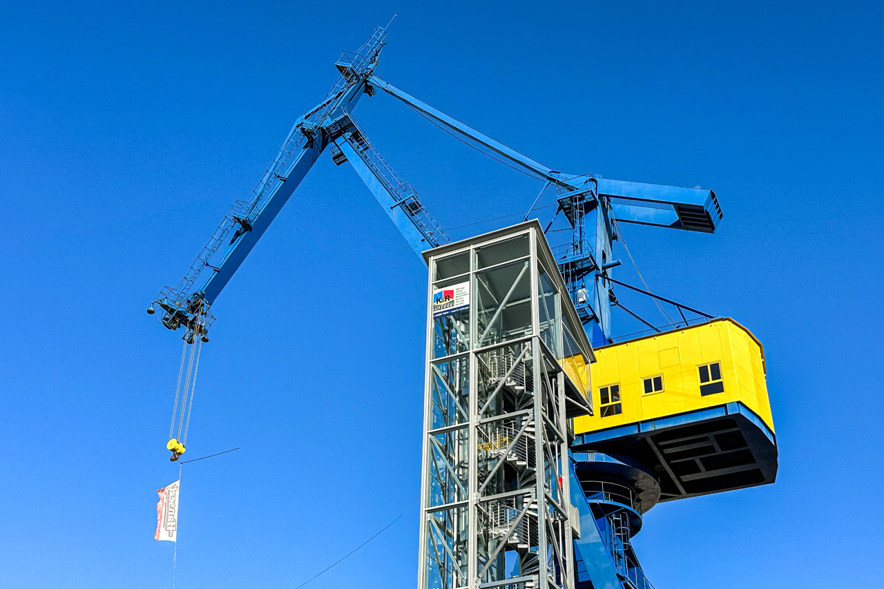 Nahaufnahme des blauen Hellingkrans von unten vor wolkenlosem blauem Himmel: Der lange Ausleger ragt schräg nach links oben, an seinem Haken hängen ein gelber Lasthaken und ein Banner. Rechts das gelbe Kranhaus mit Fenstern, davor der gläserne Turm mit Wendeltreppe und Aufzug.