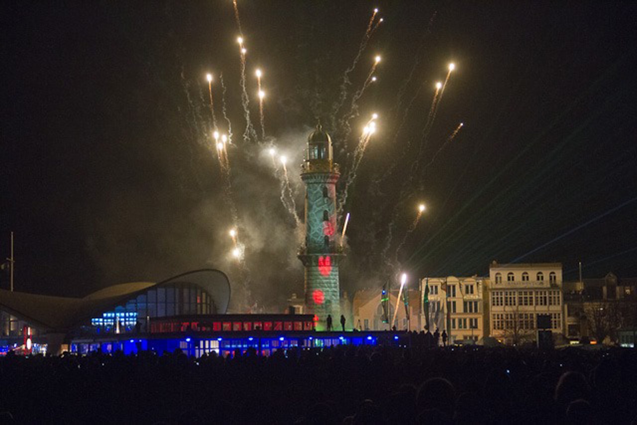 Mit wilden Mustern und Feuerwerk wird der historische Leuchtturm in Warnemünde in Szene gesetzt.
