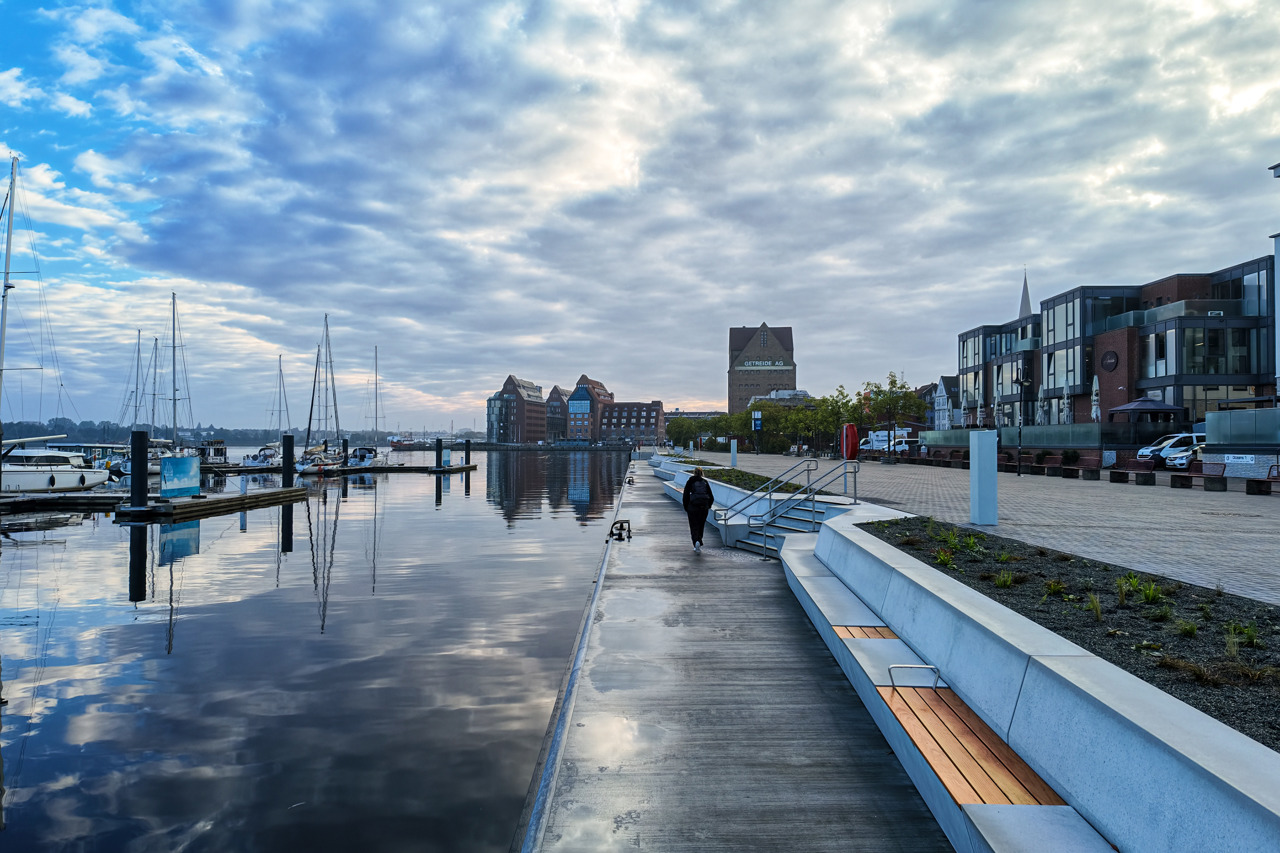 Eine Person läuft allein über die nasse Promenade am Wasser. Rechts verlaufen lange helle Sitzstufen aus Beton mit eingelassenen Holzbänken, dahinter ein frisch bepflanztes Beet und der gepflasterte Platz vor den Restaurants. Links liegen Segelboote in der Marina, im glatten Wasser spiegelt sich ein bewölkter Himmel. Im Hintergrund stehen die backsteinroten Speichergebäude der Silohalbinsel, darunter der Speicher mit der Aufschrift „Getreide AG“.