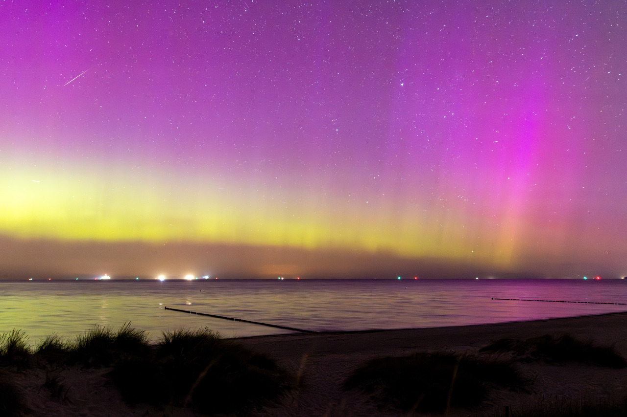 Polarlicht über der Ostsee vor Warnemünde: Direkt über dem Horizont liegt ein gelbgrünes Leuchtband, darüber färbt sich der gesamte Himmel kräftig magenta, durchzogen von senkrechten Strahlen; rechts steht ein besonders heller rosa Beamer. Links oben zieht eine Perseiden-Sternschnuppe einen feinen weißen Strich. Die spiegelglatte See wirft die Farben zurück, am Horizont die Lichter einzelner Schiffe auf Reede. Vorn dunkle Dünen mit Strandhafer, zwei Buhnenreihen ragen ins Wasser.