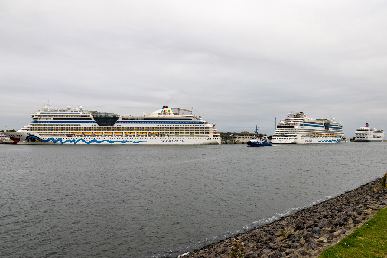 Drei Kreuzfahrtschiffe in Warnemünde: links liegt die AIDAmar mit Kussmund-Bemalung längsseits am Terminal P8, daneben rechts die AIDAluna mit dem Heck zur Kamera an P7, ganz rechts an P1-4 die Ambition mit magentafarbenem Schornstein. Ein blauer Schlepper fährt im Vordergrund durch den Seekanal, der Himmel ist grau bewölkt.