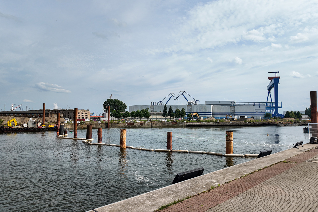 Blick auf das Werftbecken in Warnemünde. Im Hintergrund sind Hallen und der blaue Bockkahn des Marinearsenals zu sehen. Links alte Gebäude der ehemaligen Warnow Werft und ein gelber Bagger auf einer Schwimmplattform. Im Vordergrund sieht man quer durchs Bild den Schlickvorhang.