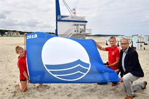 Die Rettungsschwimmer Alexandra Sievert und Arian Baume von der DRK-Wasserwacht präsentierten mit Tourismusdirektor Matthias Fromm die „Blaue Flagge“ 2018. Die Seebäder der Hansestadt Rostock wurden zum 22. Mal mit dem internationalen Umweltsiegel ausgezeichnet. (Foto: Joachim Kloock/TZRW)